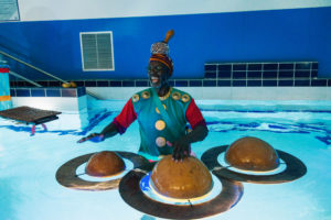 Performer in the hydrotherapy pool in an elaborate costume, playing a floating instrument