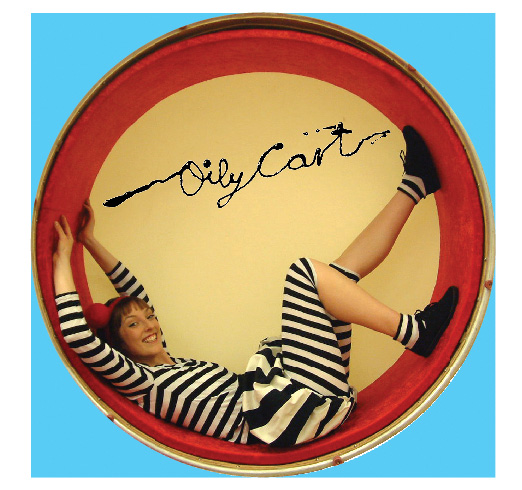 Circular image with blue border. Performer in black and white striped costume lays inside a circular drum. The Oily Cart logo is above her.
