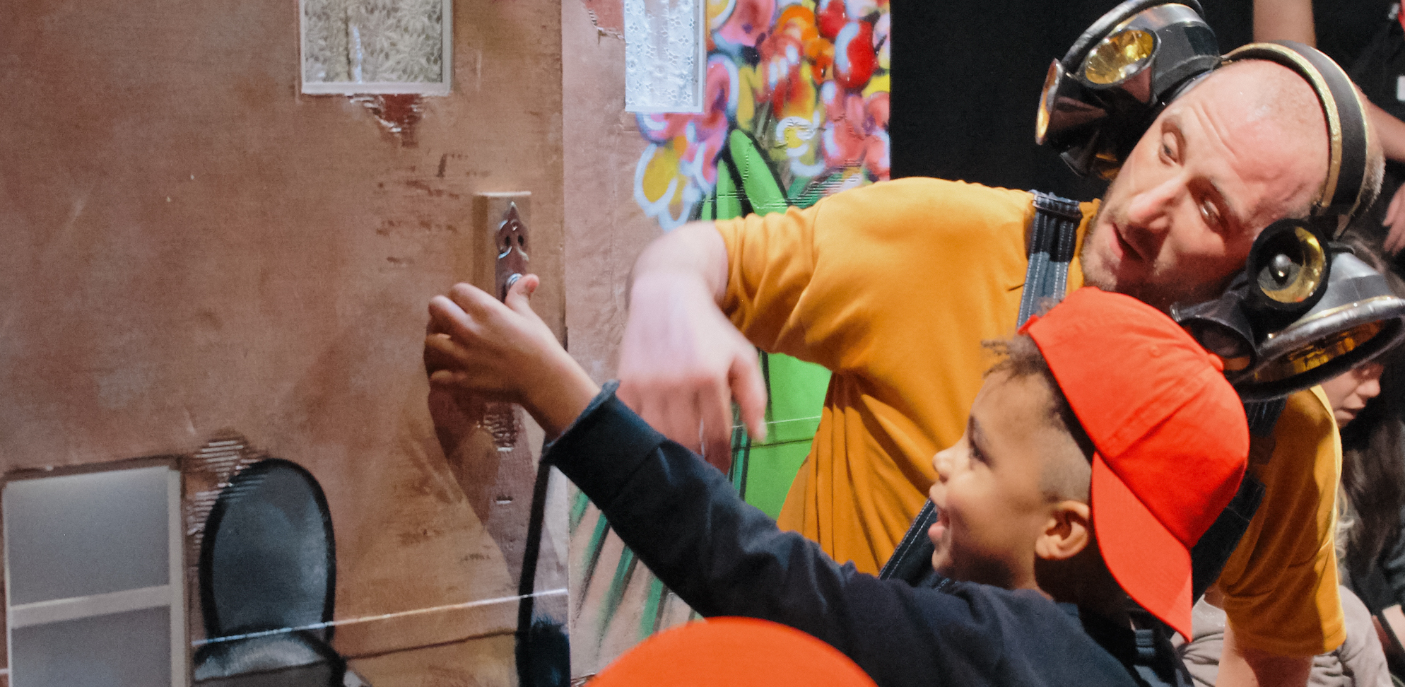 Young audience member wearing backwards orange cap presses a doorbell in the Hippity Hop Town. A performer wearing speaker headphones watches him. Photo from Hippity Hop, Credit Suzi Corker