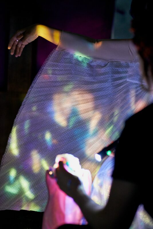 Performer's outstretched arm with pleated, winged white sleeve. In the foreground is the silhouette of a figure shining a torch on sparkly material which is reflecting colourful patterned lights onto the sleeve.