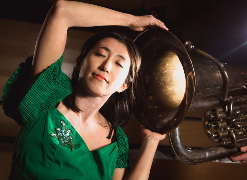 Performer listening to a brass instrument being blow directly into her ear, a flowing light visible from inside its bell.