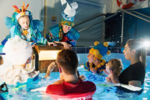 Audience in a hydrotherapy pool with performers in elaborate blue costumes, looking with awe as a treasure chest is opened