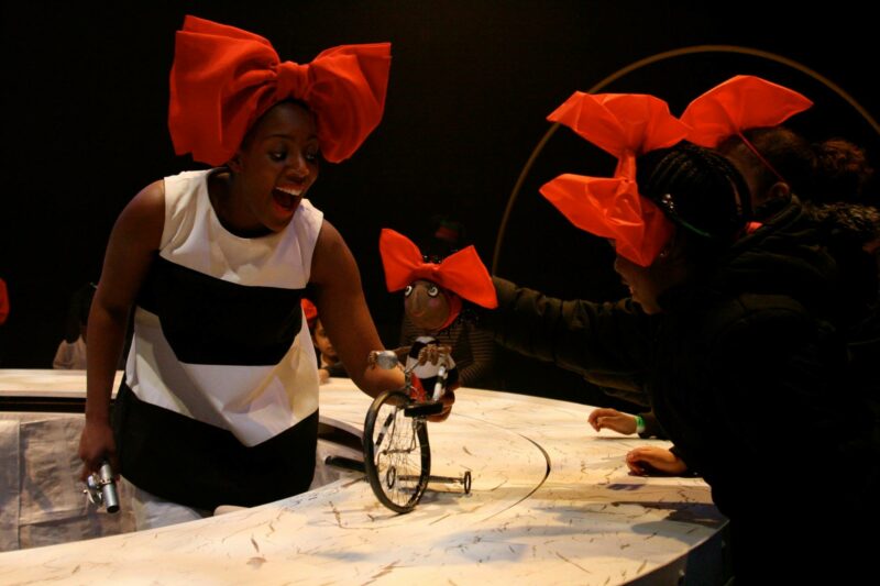 Performer in black and white costume and big red bow puppeteers an identical puppet on a bicycle, while two young audience members wearing big red bows watch.