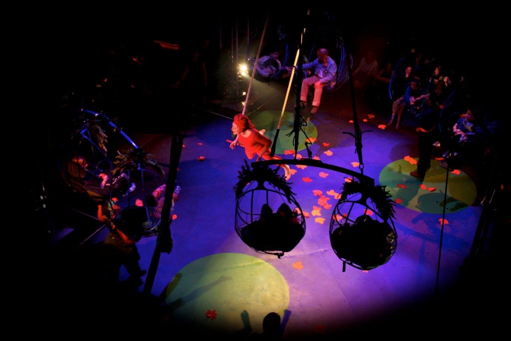 An aerial performer wearing a red costume flies through the air. The audience sit in specially-created egg-shaped seats that have been lifted into the air to surround the aerial performer.