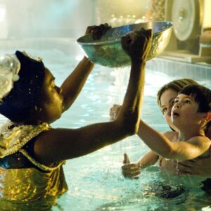Performer and audience member in a pool. The performer is holding up a large silver bowl with tiny holes in it. The child is looking up with wonder and feeling the shower of water pouring through. Photo from Pool Piece, credit Patrick Baldwin