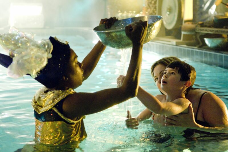 Performer and audience member in a pool. The performer is holding up a large silver bowl with tiny holes in it. The child is looking up with wonder and feeling the shower of water pouring through. Photo from Pool Piece, credit Patrick Baldwin