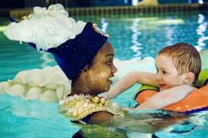 Performer wearing a hat with a cascade of bubbles interacting with a young audience member in a pool. Photo from Pool Piece, credit Patrick Baldwin