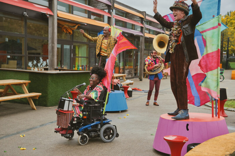 Four members of the band performing in a playground. Two of the band members stand on colourful podiums with their arms raised in the air. The singer is in a wheelchair infront of them, and a sousaphone player behind them. They are all wearing colourful festival costumes and smiling at the audience who are out of shot.