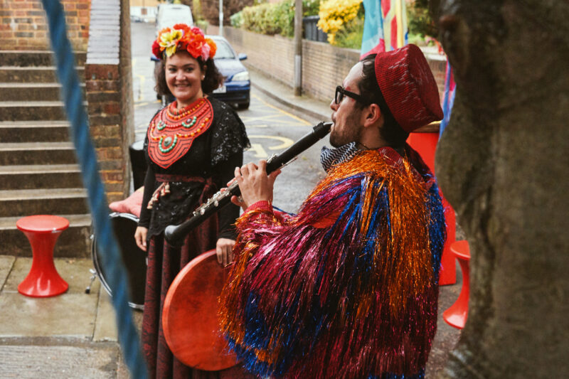 Two members of the Jamboree band are performing under a tree in a residential street. A female singer in a dark top and skirt with flowery head dress and orange beaded collar smiles at the male clarinettist who is playing in the foreground. The clarinettist is wearing a multi-coloured shiny tasselled jacket and colourful hat.