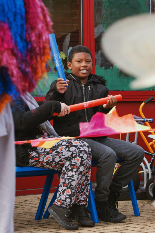 A boy sat next to a staff member in a school playground plays blue and red plastic percussion instruments. In the foreground is a musician in a colourful tasselled jacket with his back to the camera.