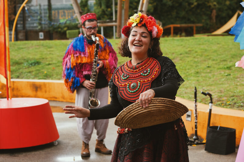 Two members of the Jamboree band are performing in a plyground. A female singer in a dark top and skirt with flowery head dress and orange beaded collar sings. She is holding a flat basket. Behind her is musician playing a bass clarinet. He is wearing a multi-coloured shiny tasselled jacket and colourful hat.