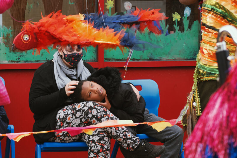 A boy sits with his head rested on a staff member's lap in a school playground. He is looking up at a colourful macaw puppet flying overhead.