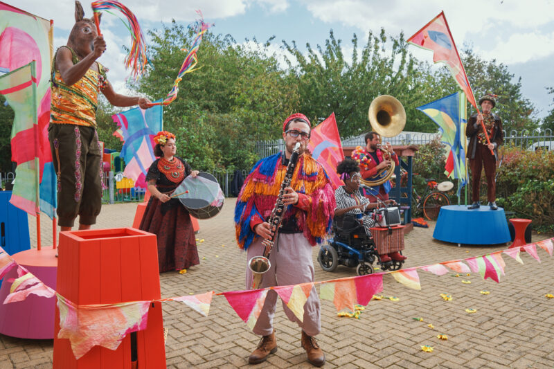 The Jamboree band, all wearing colourful costumes, perform in a playground. Colourful bunting marks out the performance space. On the left and right are the hosts standing on platforms waving flags. One is wearing a donkey mask. In the foreground is a bass clarinettist, behind him are singers and a musician playing the sousaphone.