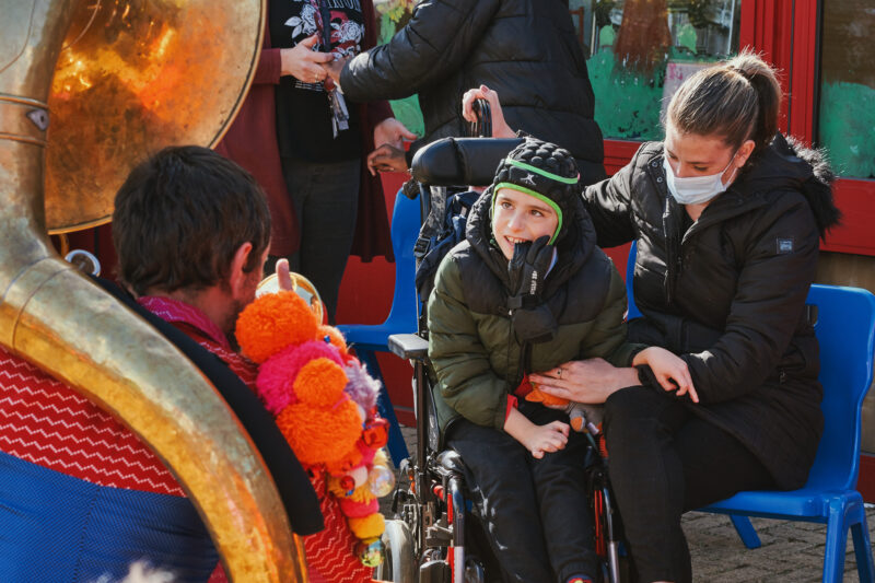 A boy sat in a wheelchair next to a staff member in a school playground, watching a musician who is playing close to them. The musician, whose back is to the camera, is kneeling. He is wearing a shiny pink and blue jacket with big colourful pom poms on the shoulder and playing a sousaphone.