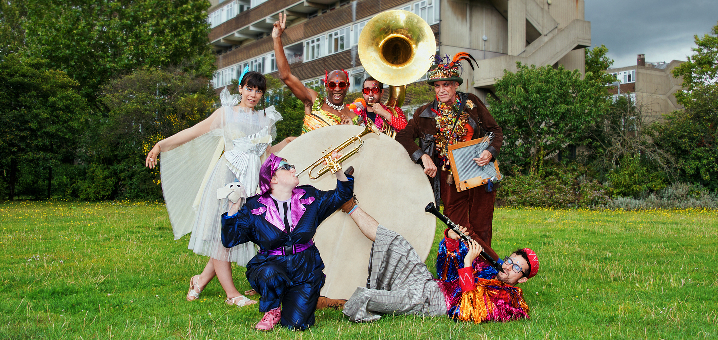 Photograph of the Jamboree band in a field. They are all wearing colourful, festival outfits and holding their instruments. They are stood around a huge drum. In the background are trees and a block of flats. Photo from Jamboree, credit Suzi Corker