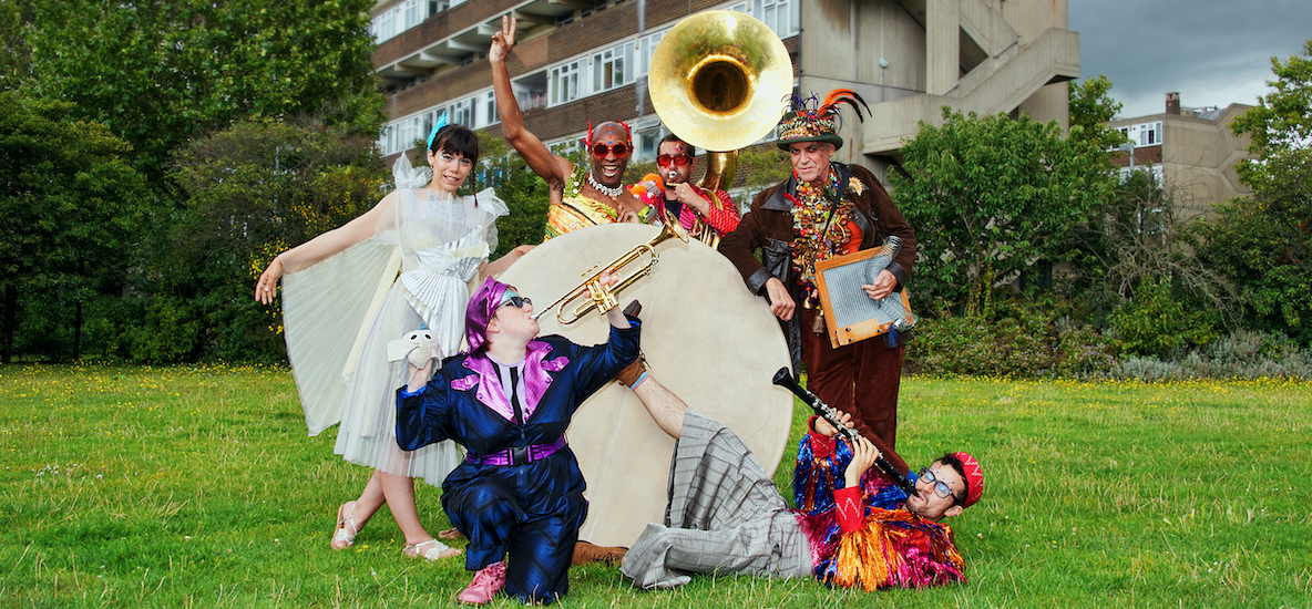 Photograph of the Jamboree band in a field. They are all wearing colourful, festival outfits and holding their instruments. They are stood around a huge drum. In the background are trees and a block of flats. Photo from Jamboree, credit Suzi Corker