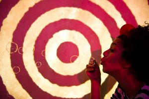 A performer blows bubbles across a background of red and white concentric circles. Photo from Drum, credit Patrick Baldwin