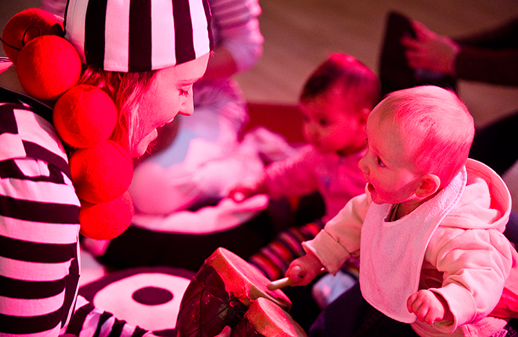 Performer in black and white striped hat interacts with a baby beating a drum. Photo from Drum, credit Patrick Baldwin