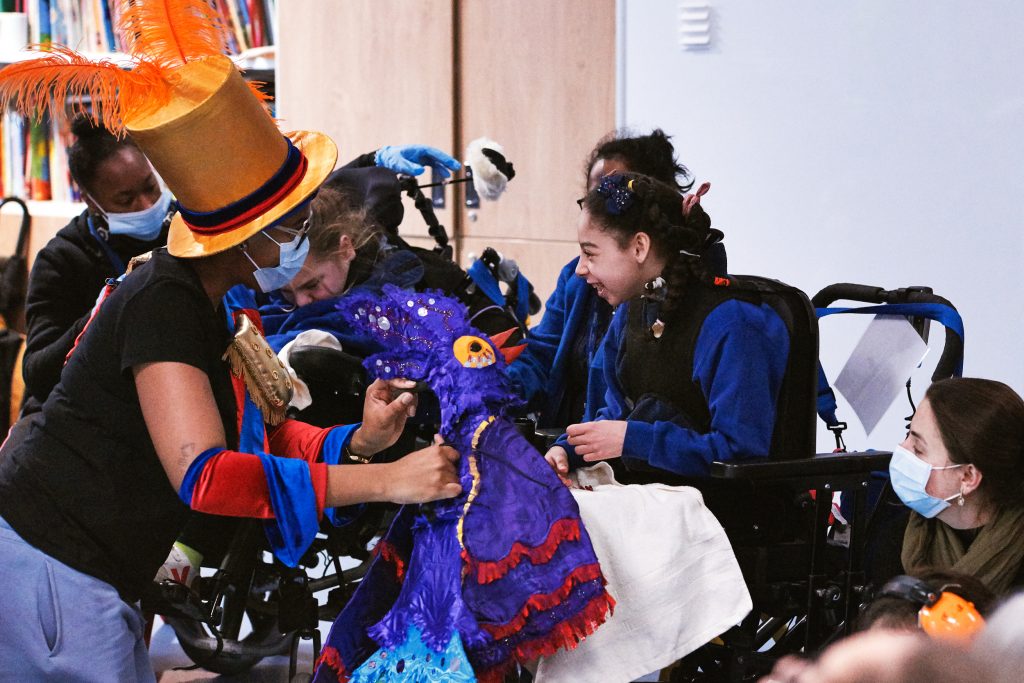 Teacher wears an orange top hat with big orange feathers and holds a purple parrot puppet. She interacts with a smiling student in a wheelchair. They are surrounded by other staff and students.