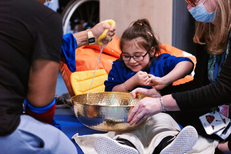 A smiling child sits on the ground with her hands clasped. She is looking into a copper bowl that is held in front of her. A performer is squeezing water from a sponge into the bowl
