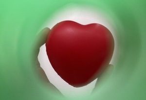 Photograph looking through a green straw. At the other end is a hand holding a red heart. Photo from Something Love, Credit: Franki Ayres
