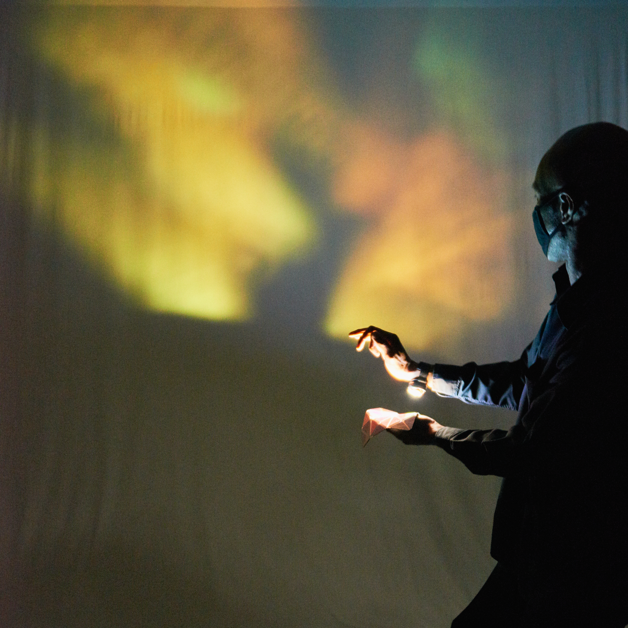 A Black man shines his wrist torch onto a piece of shiny material with mirror triangles. This causes colourful light patterns on the wall behind. Photo from Space to Be, credit Suzi Corker