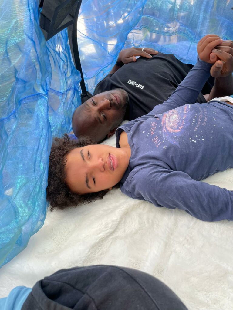 A parent and child lie holding hands on a soft white rug in a tent with shimmery blue and green gauze walls