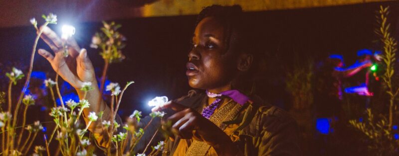 A performer with small bright lights attached to a finger on each hand. They holds their hands aloft, near branches with leaves and flower buds.