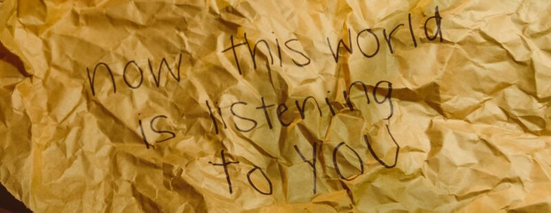 Scrunched brown paper laid flat on a table. In black felt pen is written Now this world is listening to you.