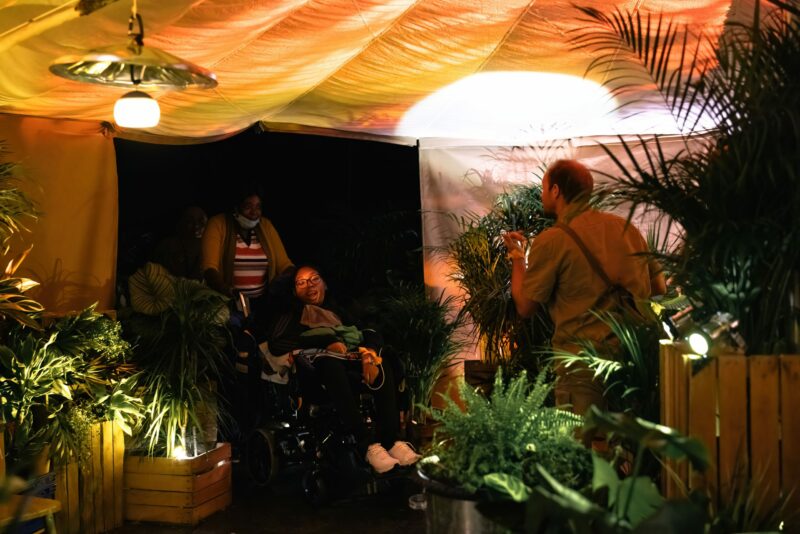 A performer with a torch guides audience members into a large tent. It is filled with plants in wooden and plastic crates. A metal bin lid with a bulb hanging from it hangs overhead.