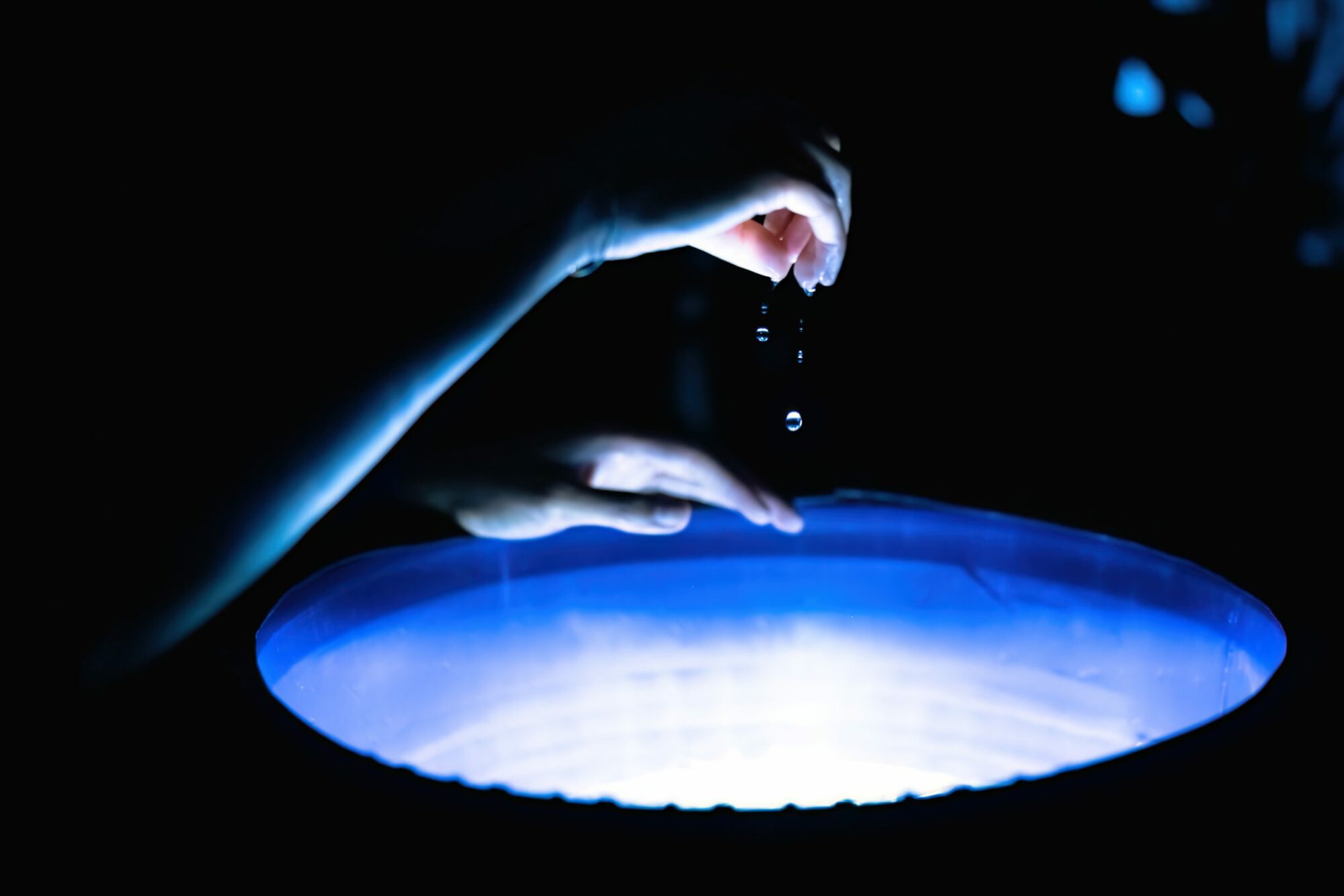 A circular bin filled with water in a dark space. A light underneath illuminates the water. Water droplets fall from a hand held above it.