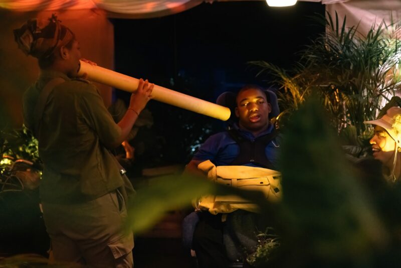 A photo taken between plant leaves. A performer holds up a long tube, making sounds, towards the ear of a young audience member. An adult sat alongside them looks up smiling. They are surrounded by plants.