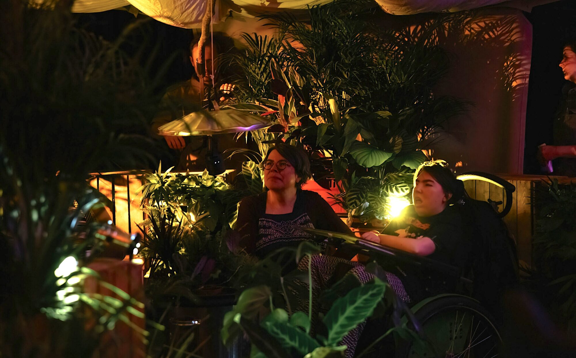 A young woman with short brown hair sitting in her wheelchair. A middle aged woman with short grey hair and glasses sits next to her. They are inside the When the World Turns performance space, with a billowing tent overhead and surrounded with wooden crates and plants all around. There is atmospheric low lighting, illuminating their faces with a golden glow and casting shadows of the plant leaves. Hanging just above them is a metal bin lid.