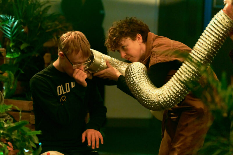 A performer holds the trunk of an elephant puppet, made from a long silver corrugated tube, to the ear of a young member.