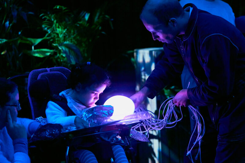 A young person looks at a jellyfish puppet held close to them by a performer. The body of the jellyfish is a light, which illuminates the young person's face with a pink glow.