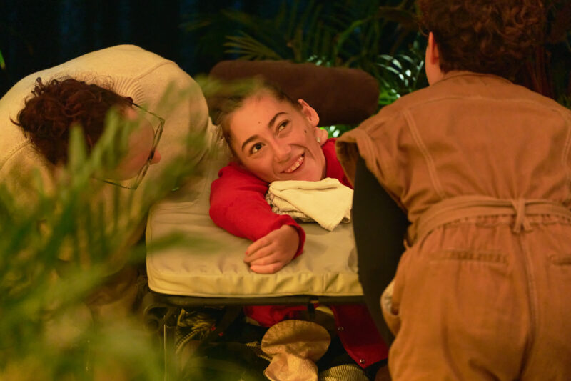 An audience member viewed through leaves. They are smiling up at a performer.