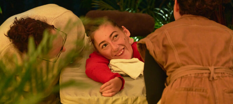 An audience member viewed through leaves. They are smiling up at a performer.