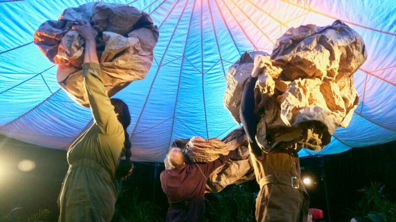 Three performers in earth-coloured overalls. They hold huge scrunched up balls of thick brown paper over their heads. A parachute tent roof overhead is lit in pinks and blues.
