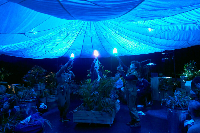 Three performers hold jellyfish puppets aloft, made of glowing lights and recycled electrical cords. The whole space is lit with a blue light, like the audience are under the sea.