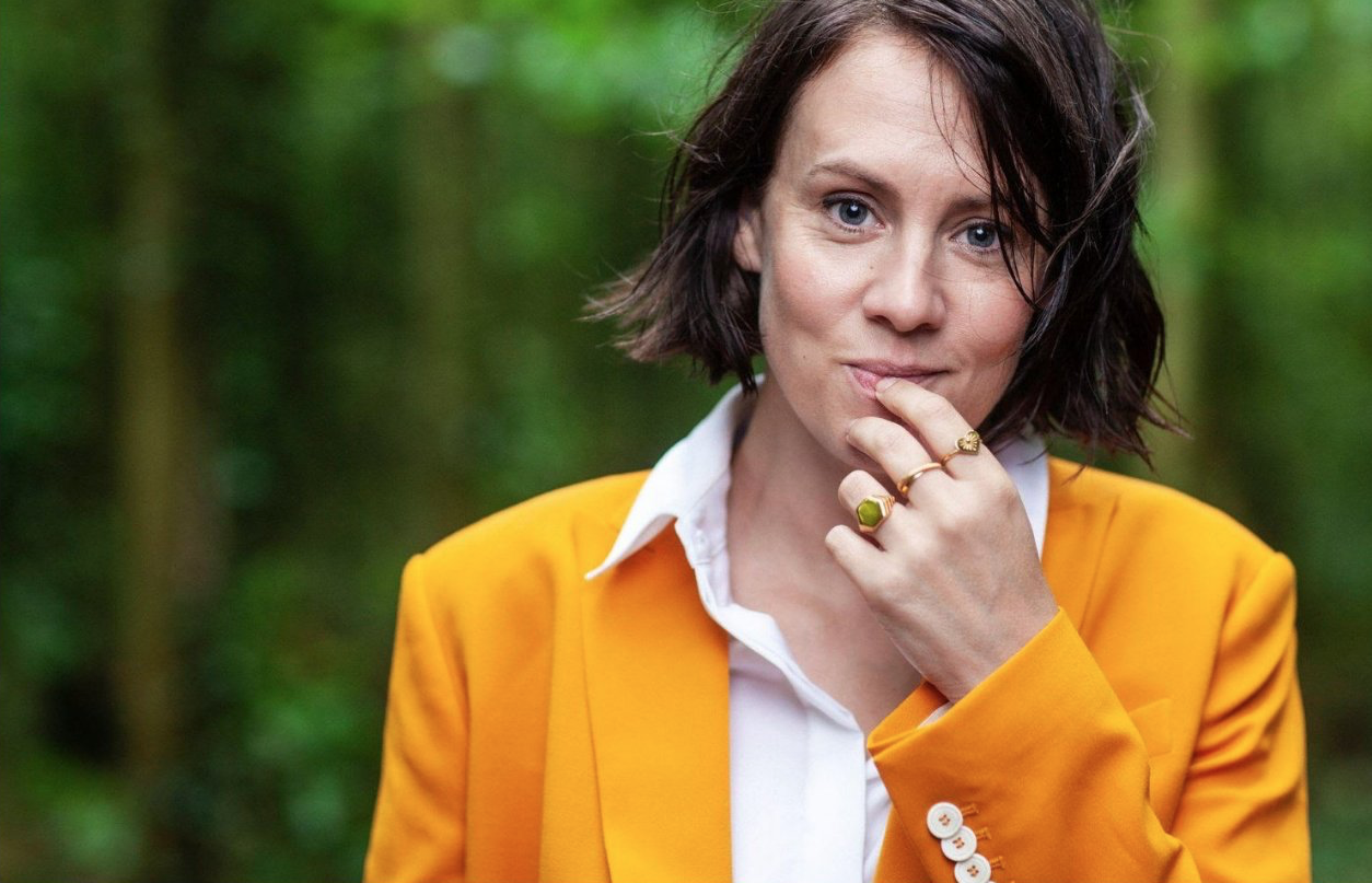 Bryony Kimmings is standing in front of a backdrop of green trees. She is wearing a yellow jacket and her left hand is lifted to her chin.