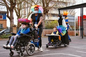 A girl and a boy in wheelchairs are pushed through a school playground by their female teachers. The girl is wearing a pink and blue straw hat. Her teacher is wearing a multicoloured umbrella hat. The boy's chair is decorated with a multicoloured netting cape. A Cart performer in a top hat decorated the orange feathers brings up the rear. All the adults are wearing medical masks.