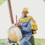 A kora musician sits on a green stool upon a cream coloured carpet decorated with light and dark green leaves. Two large decorate leaves frame his head. The musician balances the base of the kora between his knees above the floor. The kora is a large gourd with a long neck traversed by 21 strings. The man wears a light blue cordoroy jumpsuite, yellow socks and blackshoes. Around his kneck is a yellow and blue frilled collar with a matching cape attached. On his head he wears a yellow cap with three small fabric horns attached.