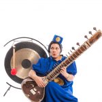 A woman sits on the floor playing a sitar. She is dressed in a bright blue jump suit and a matching bell boy hat decorated with a gold circle; the toe nails of her bare feet are painted dark blue. Behind her right shoulder stands a large bronze gong; a wooden beater with a red furry head, hangs from the gong.