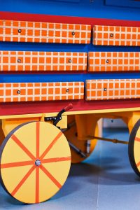 A close-up picture showing six long wooden bars (embaire bars) fixed to the side of The Cart with shiny metal screws. The bars are painted in orange and white checks. The base of the cart is painted in blue and red stripes with a yellow trim. One of The Cart's wheels painted yellow with red spokes is visible. The Cart is parked on a blue floor.