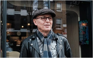 A middle aged man stands in front of a shop window with buildings reflected in the glass. He wears a black/grey tweedy cap, a black leather jackets, grey/blue check scarf and a grey and black patterned shirt. He is wearing glasses with dark frames.