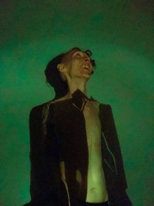 A green-tinted image of a smiling man in a dark jacket open over a bare chest. His shape makes a distinct dark shadow against a green background.