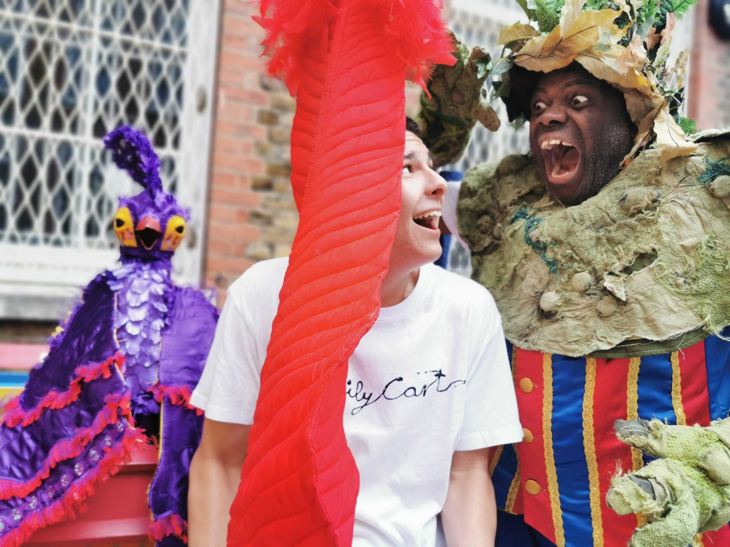 A performer playing the part of a scary troll bares his teeth and snarls at a young man. The young man's face is partly obscured by an enormous red fabric feather. He looks up at the troll in troll in mock alarm. He wears a white t-shirt with the words Oily Cart written across the front in black. The troll wears a red and blue striped waistcoat with a pale khaki coloured colour and matching hat decorated with leaves. Behind the young man, a purple fabric bird looks on behind the young man.