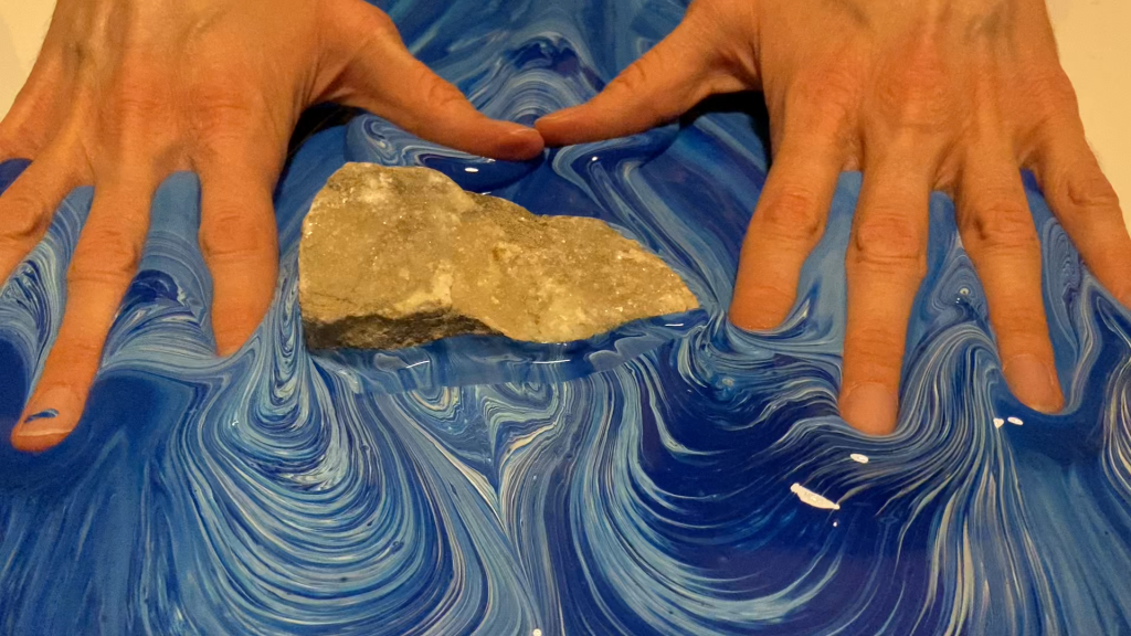 Hands with fingers splayed dip into a swirl of blue and white paint. A piece of sandstone sits in the paint in the space between the two hands where the tips of the thumbs meet.
