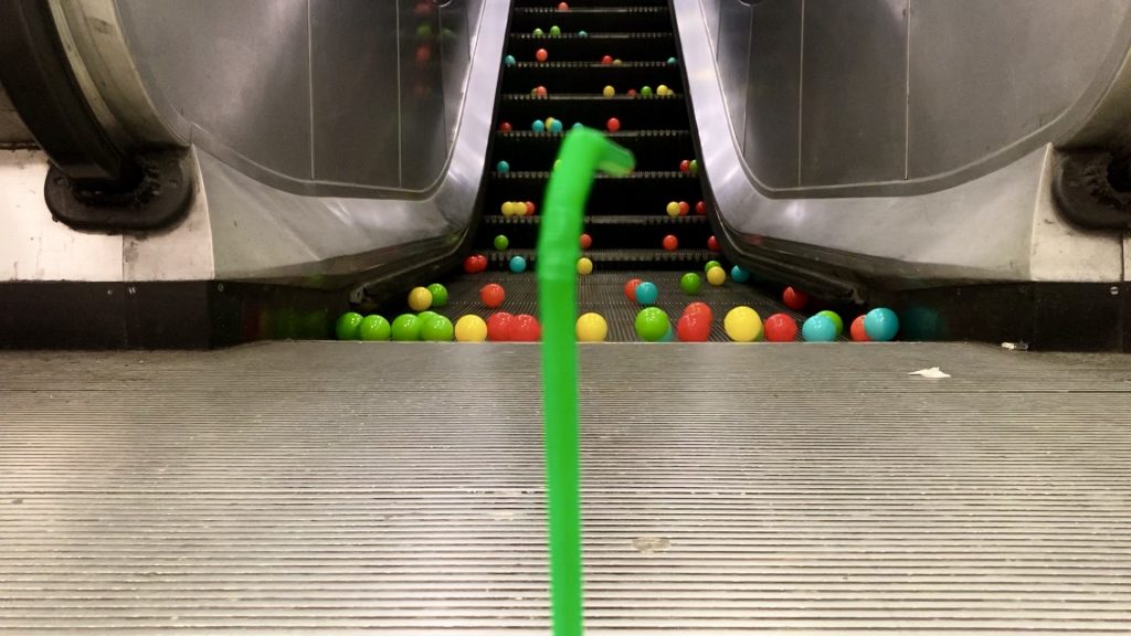 An upright green plastic straw at the bottom of an escalator. Green, red, yellow and turquoise plastic balls are scattered over the steps of the escalator.