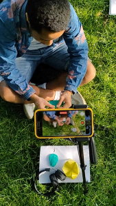 A young person sits crosslegged on the grass wearing blue denim shorts and jacket embroidered with pink flowers. In front of them on the grass there is a sheet of white paper with a folded yellow paper shape, black headphones, and a lump of turquoise "squish" (like plasticine). The scene is filmed by a phone camera suspended above the paper.